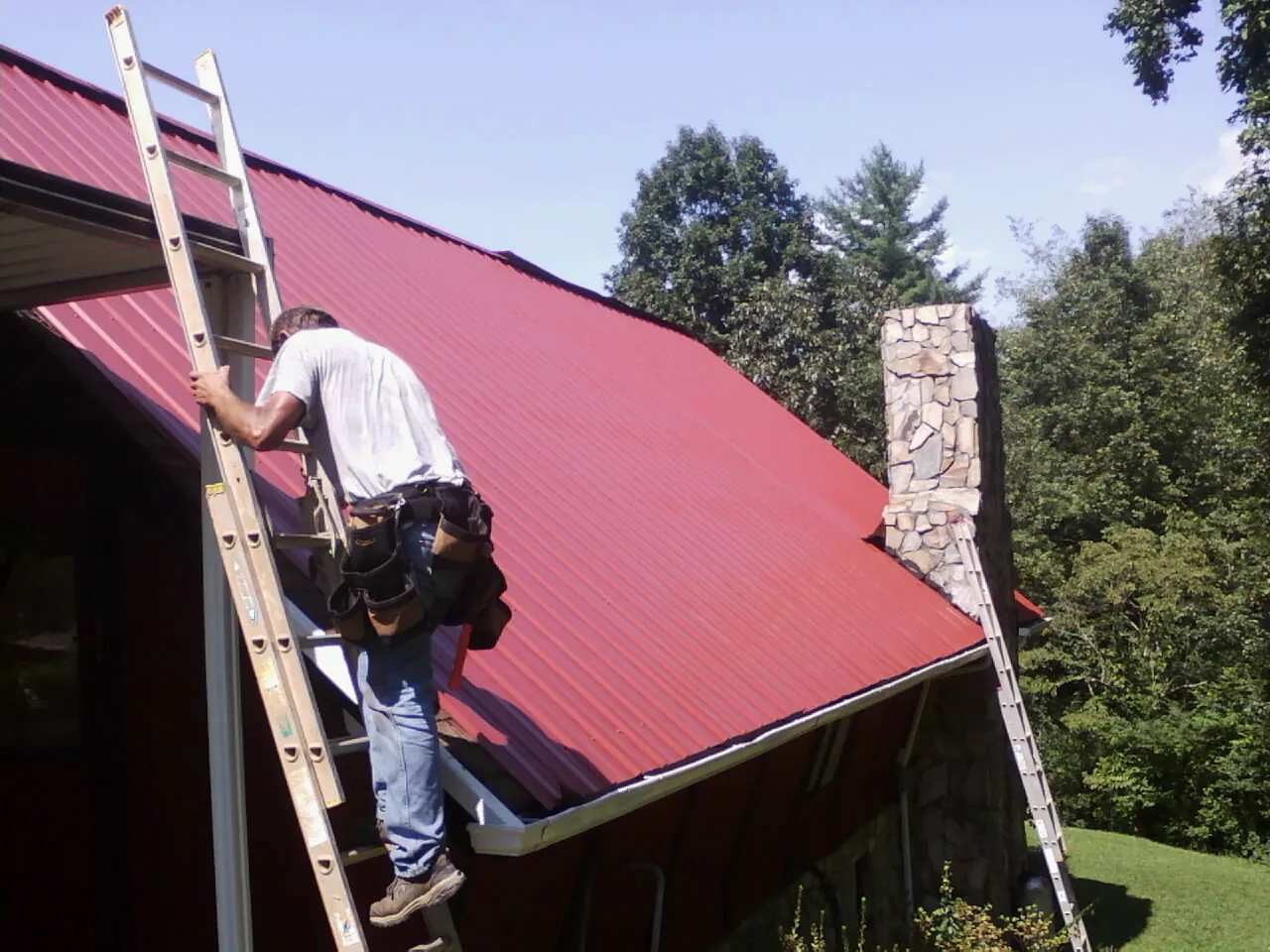 Roofer on a ladder beside a red metal roof and stone chimney — Soffit & Fascia Repair by Roofing Brothers in Garden Valley